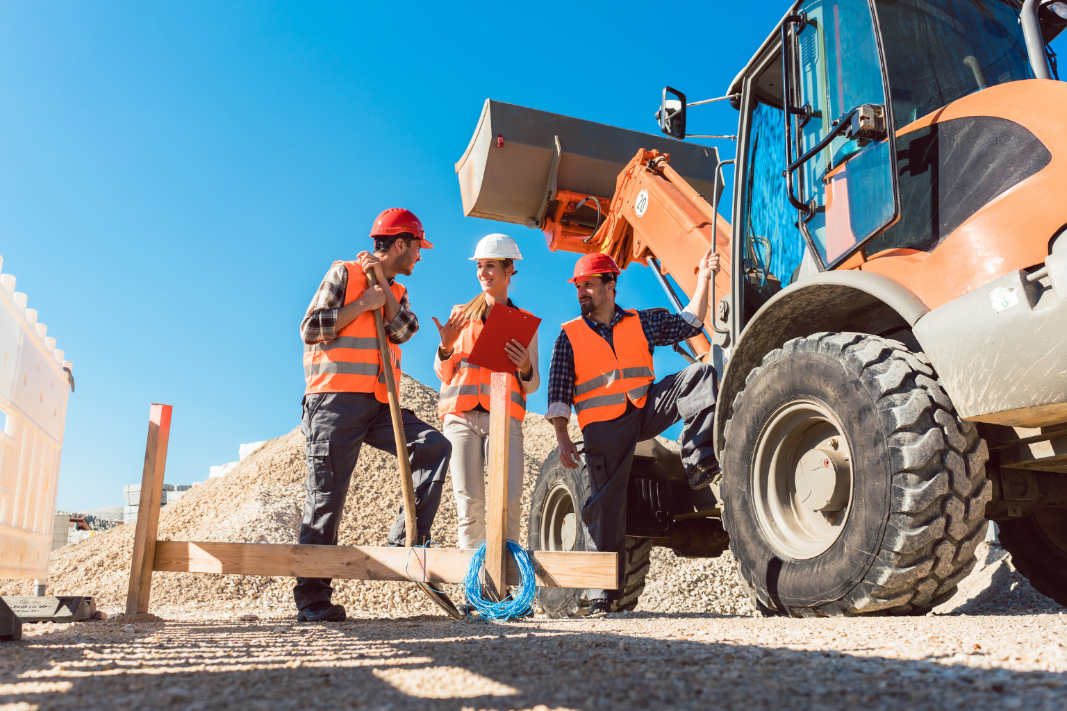 Three construction workers in high-visibility gear talking near heavy equipment.